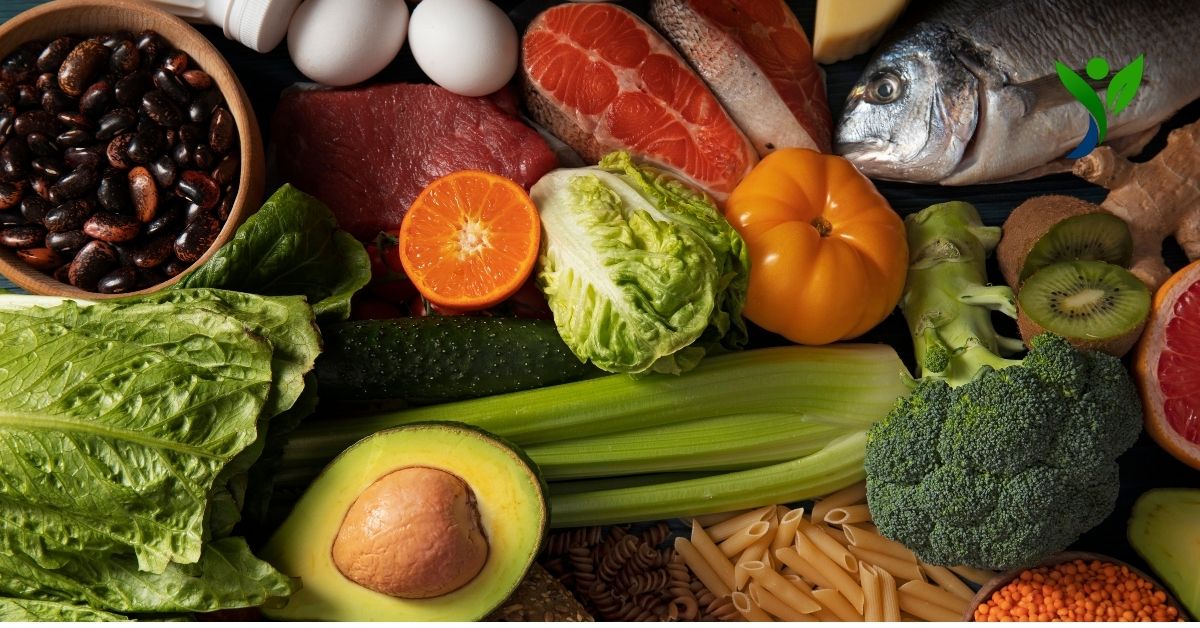 A variety of nutrition-rich foods, including leafy greens, avocado, beans, eggs, lean meat, salmon, whole fish, tomato, orange, broccoli, kiwi, celery, pasta, and whole grains, displayed together.